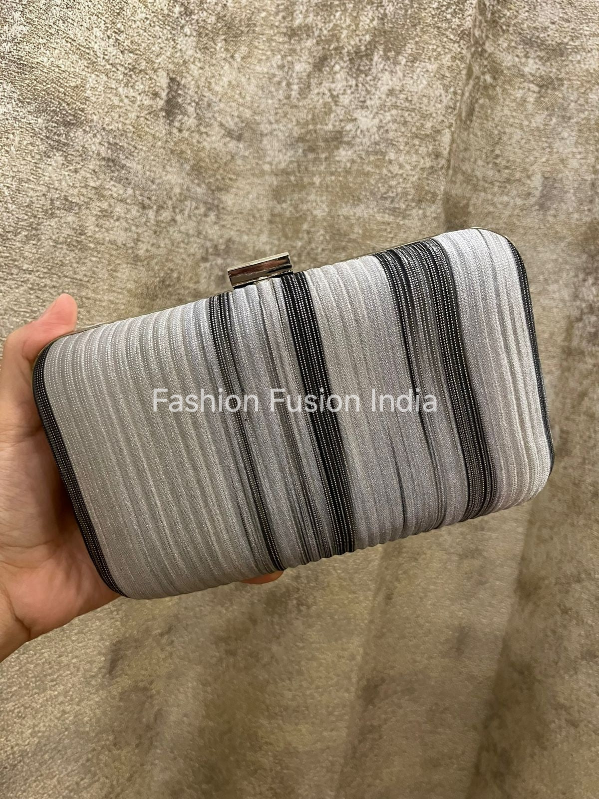 Striped clutch held by a hand on a textured surface with 'Fashion Fusion India' branding.