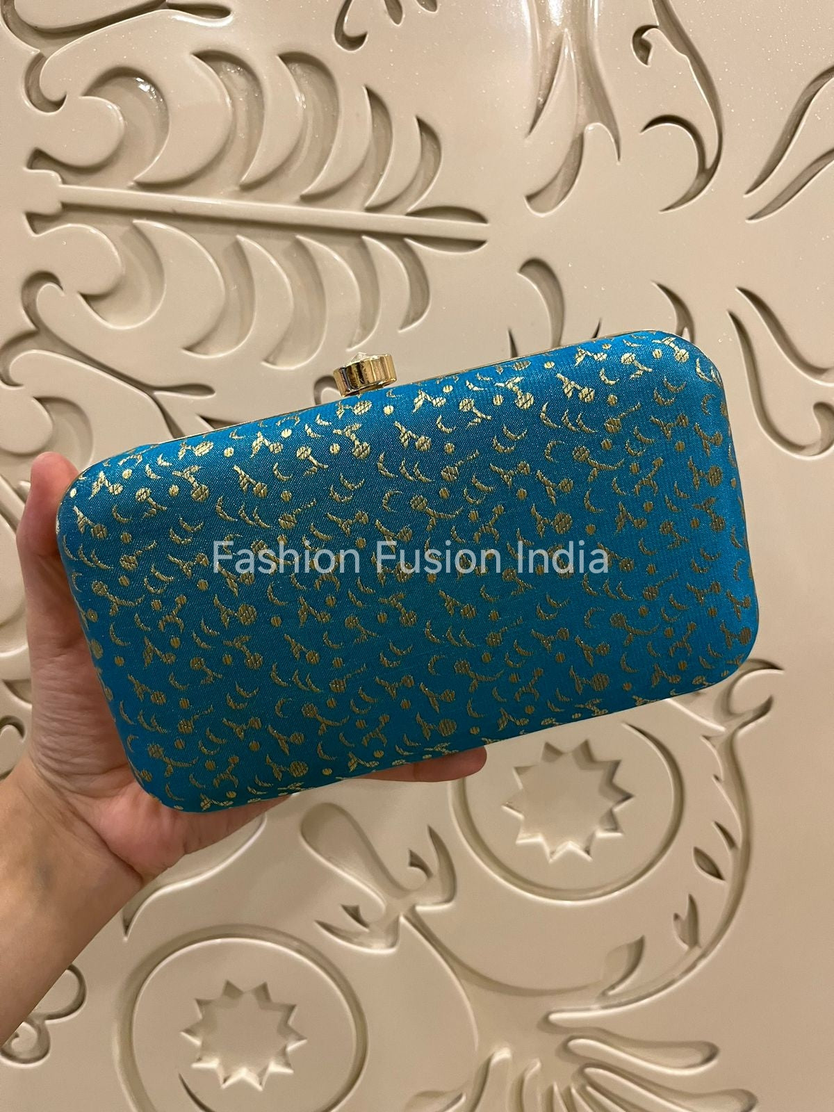 Blue clutch with gold patterns held by a hand against an ornate beige background, branded 'Fashion Fusion India'.