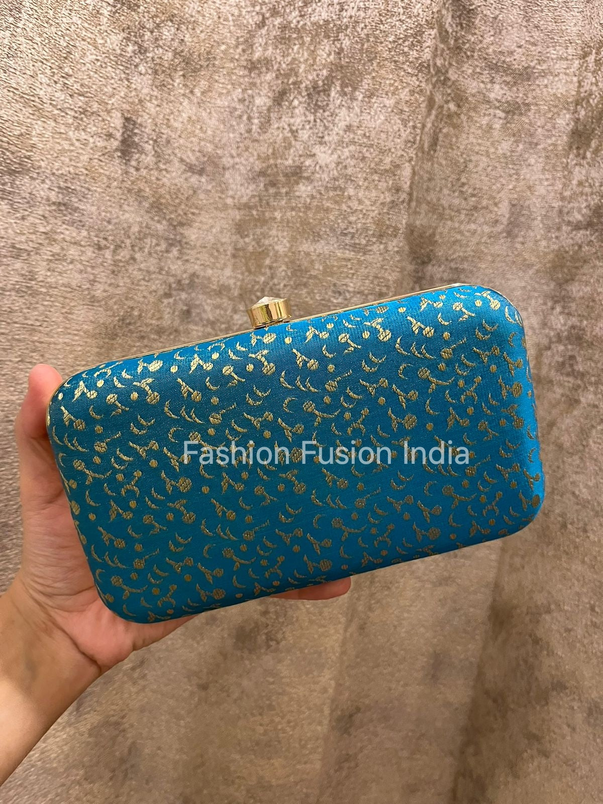 Blue clutch with gold patterns held by a hand against a textured brown background, branded 'Fashion Fusion India'.