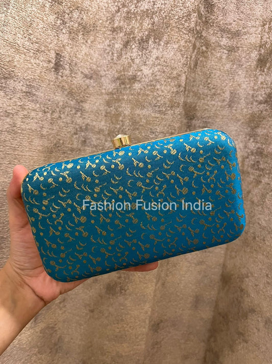 Blue clutch with gold patterns held by a hand against a textured brown background, branded 'Fashion Fusion India'.