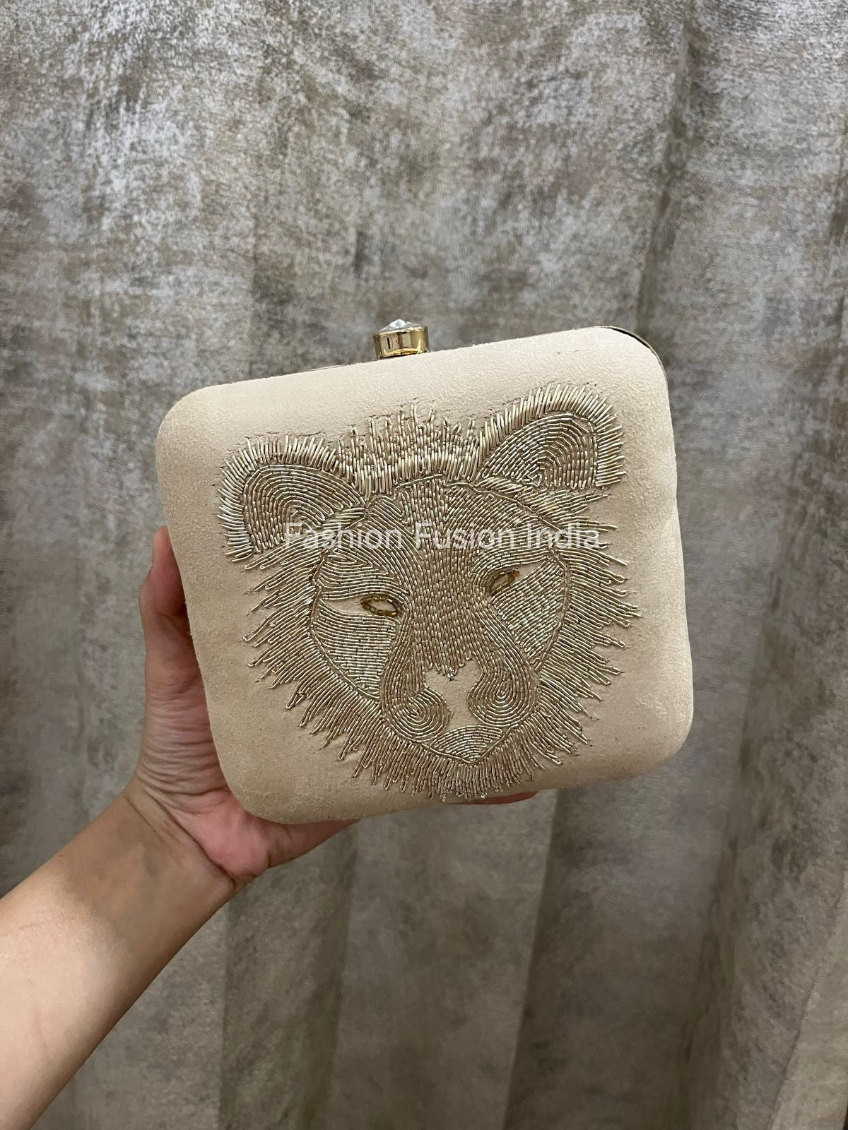 Beige clutch bag with embroidered lion design held by a hand against a textured gray background.