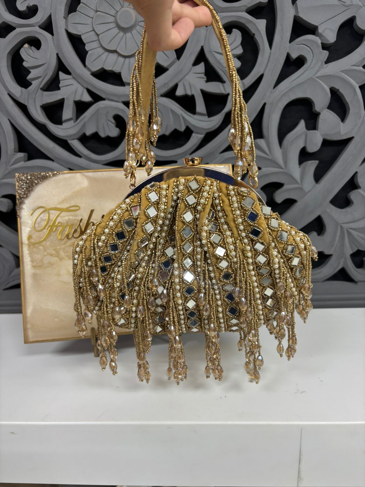 Gold beaded handbag with intricate design held by a hand against a decorative background