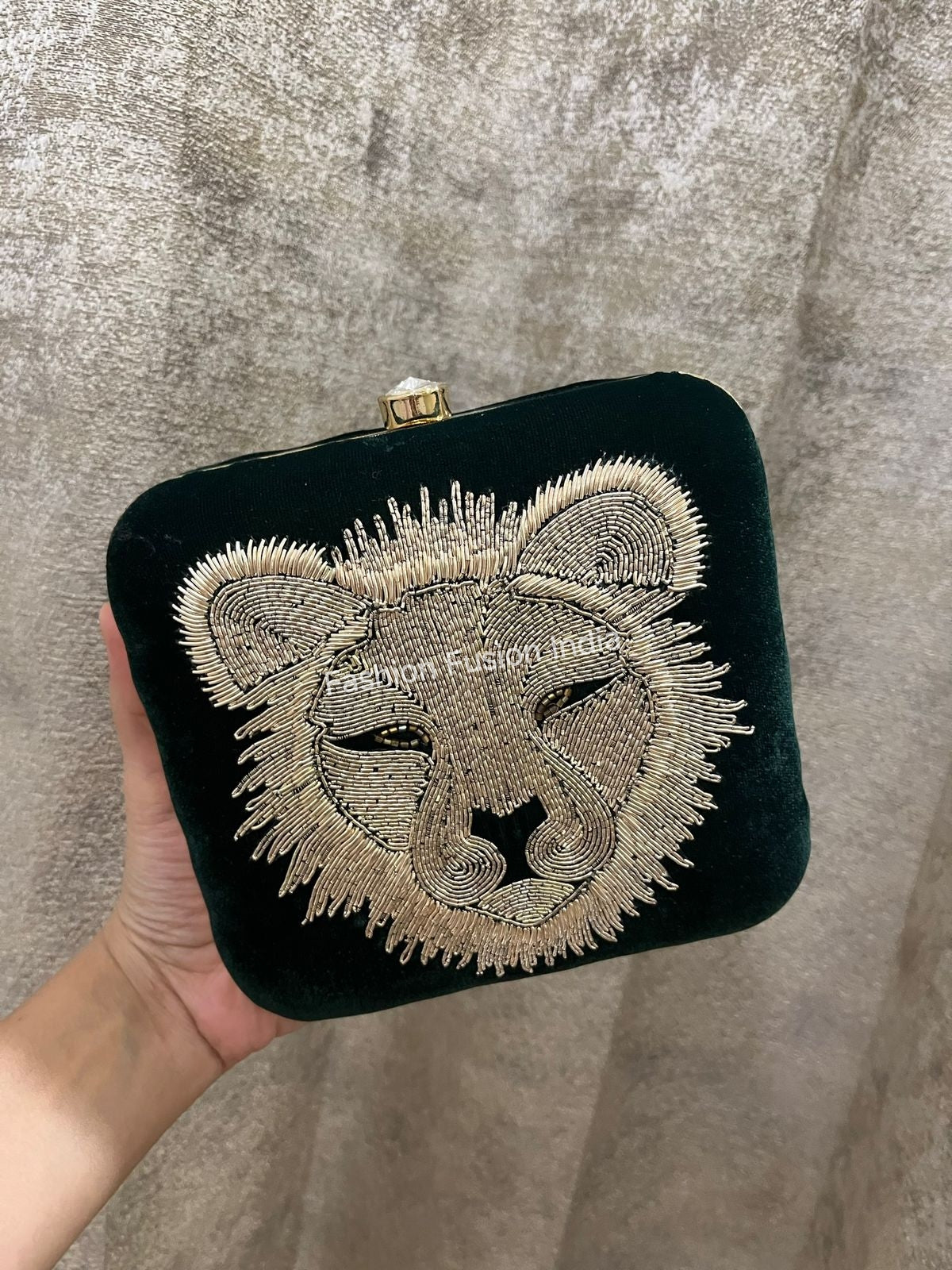 A green clutch with lion head design held by a hand against a textured gray background