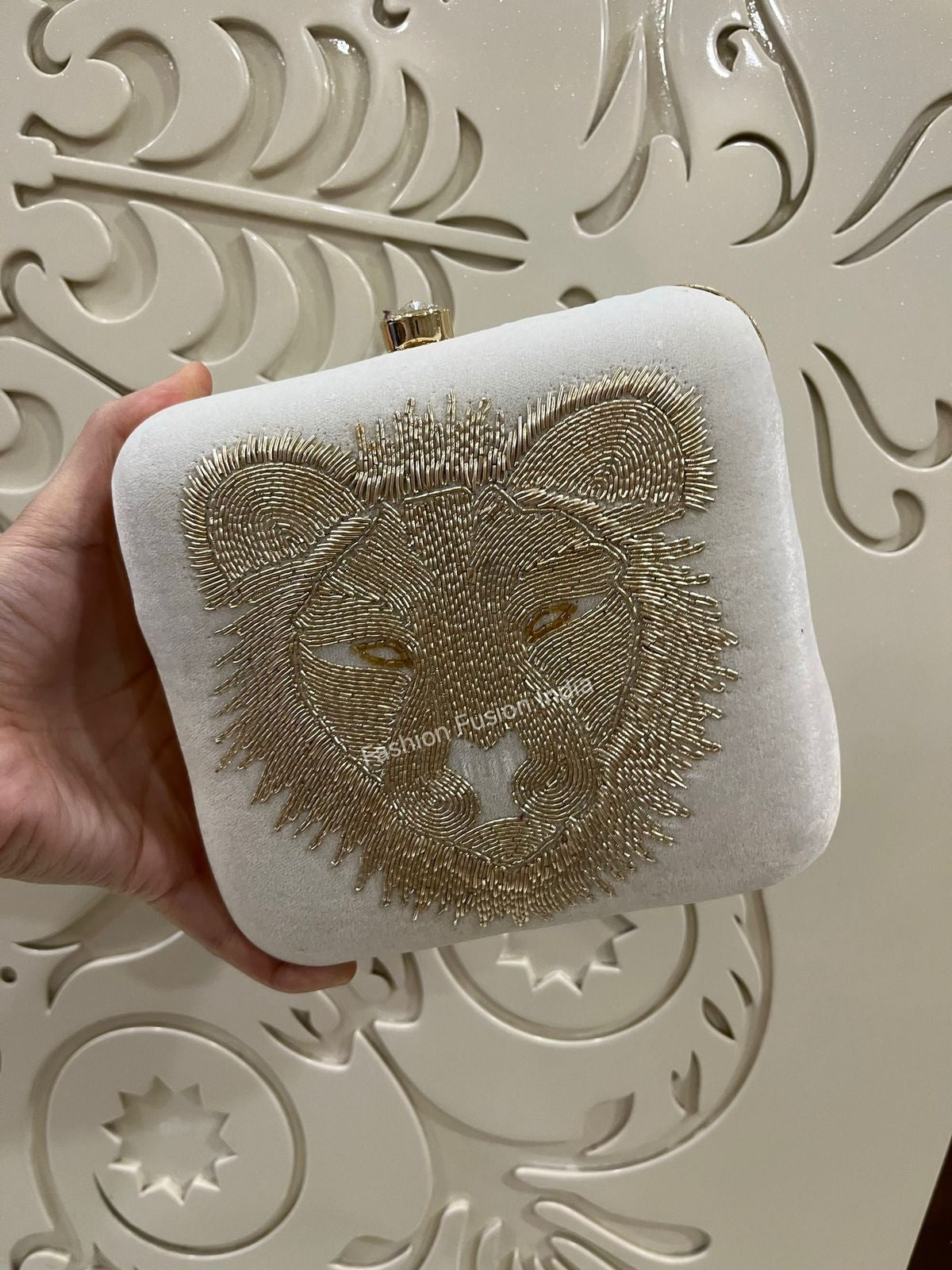 Hand holding a white clutch with gold lion embroidery against an ornate white background